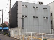 Small gray three-story building with narrow windows and fenced parking area on a quiet street, utility pole and wires alongside.