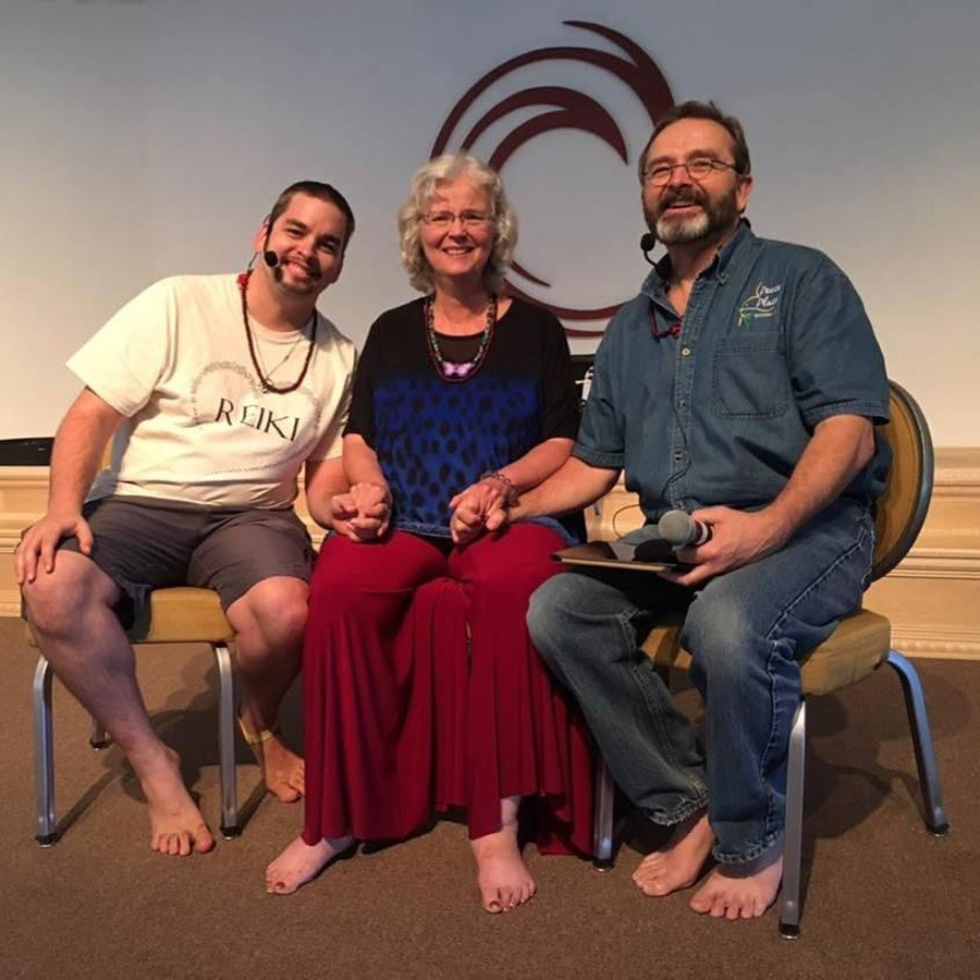 Three barefoot adults sit on chairs, smiling and holding hands, with microphones and a circular design on the wall behind them.