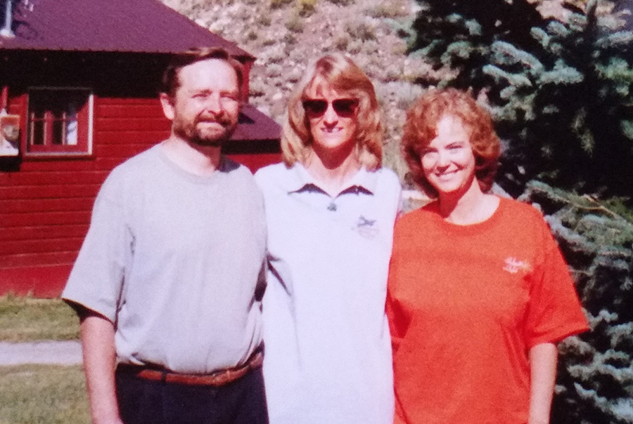 Michael Baird, Carolyn Musial and Laurelle Gaia standing outdoors, smiling, with a red building and a pine tree in the background on a sunny day.