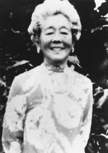 Black-and-white photo of Hawayo Takata smiling, wearing a patterned dress, standing outdoors in front of dark foliage.