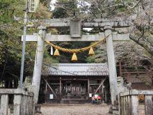 Stone torii gate with hanging rope and bells leading to a traditional wooden Shinto shrine surrounded by trees and forest path.