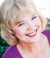 Smiling woman with short blonde hair, wearing a purple top, outdoors with greenery in the background.
