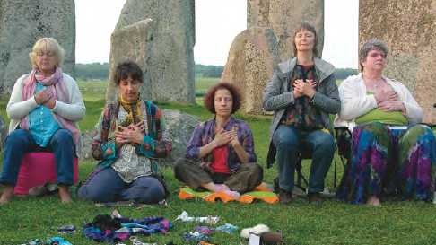 Five people seated outdoors with hands on their hearts, meditating together among standing stone monuments on a grassy field at Stonehenge.