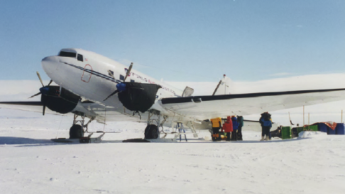 A vintage propeller airplane parked on a snowy airfield at the South Pole with people and equipment gathered beneath its wing.